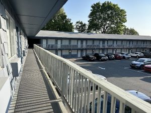 Balcony walkway along a two-story motel with blue-gray siding, railing shadows, and a parking lot with cars at Acadia National Park.