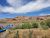 Blue inflatable rafts with paddlers drift along a muddy river, red rock cliffs rise in Arches National Park under a bright blue sky.
