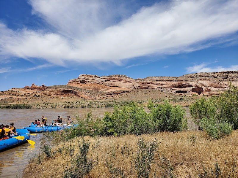 Blue inflatable rafts with paddlers drift along a muddy river, red rock cliffs rise in Arches National Park under a bright blue sky.