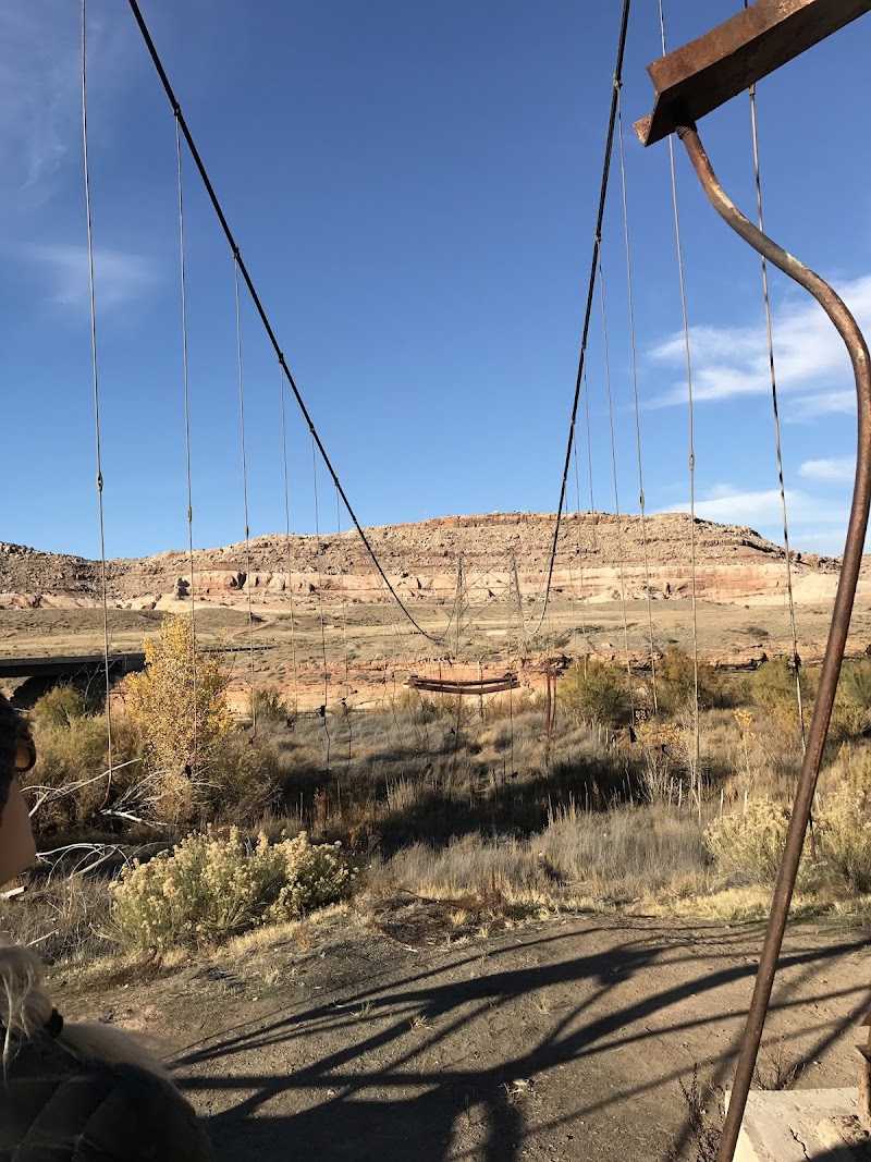 Rusted suspension bridge frame and cables over a desert campground in Arches National Park, with red rock mesas and clear blue sky.