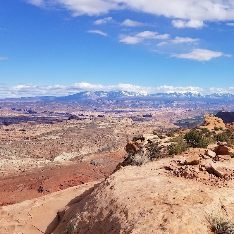 Arches National Park overlooks a vast red rock desert with distant snow-capped mountains and rock cairns along the cliff.