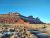 Snow-dusted red sandstone ridges rise over a desert parking area with wooden fences, dry grasses, and parked cars beneath a bright blue Arches National Park sky.