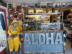 Inside a seaside cafe in Acadia National Park, a corrugated counter with a large ‘ALOHA’ sign, a yellow-raincoat mannequin, pineapple decorations, and a glass dessert display.