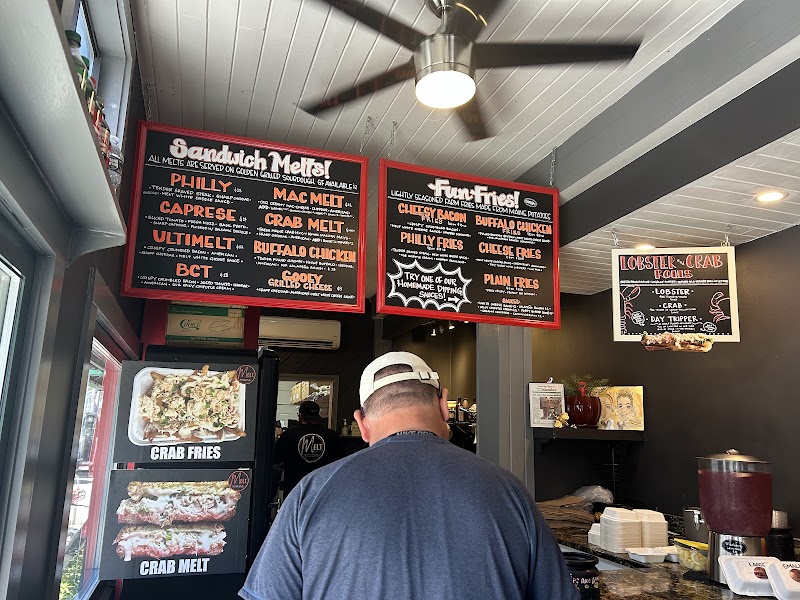 Inside a fast-casual counter at Acadia National Park, a man in a cap faces red-framed sandwich menus above the counter.