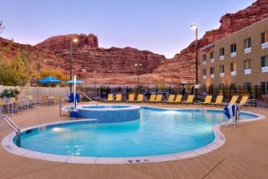 Outdoor swimming pool with lounge chairs and blue umbrellas, set against red rock cliffs at Arches National Park.