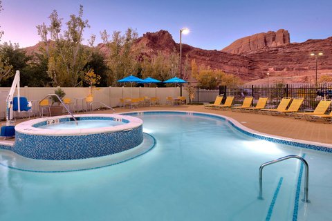 Pool area with a round raised hot tub, blue-tiled edge, yellow lounge chairs, blue umbrellas, and the desert cliffs of Arches National Park in the background.