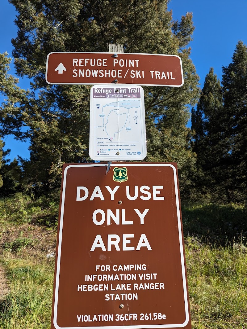Refuge Point Trailhead signs and a map board, with a 'Day Use Only Area' sign, set among pines under a blue sky in Yellowstone National Park.