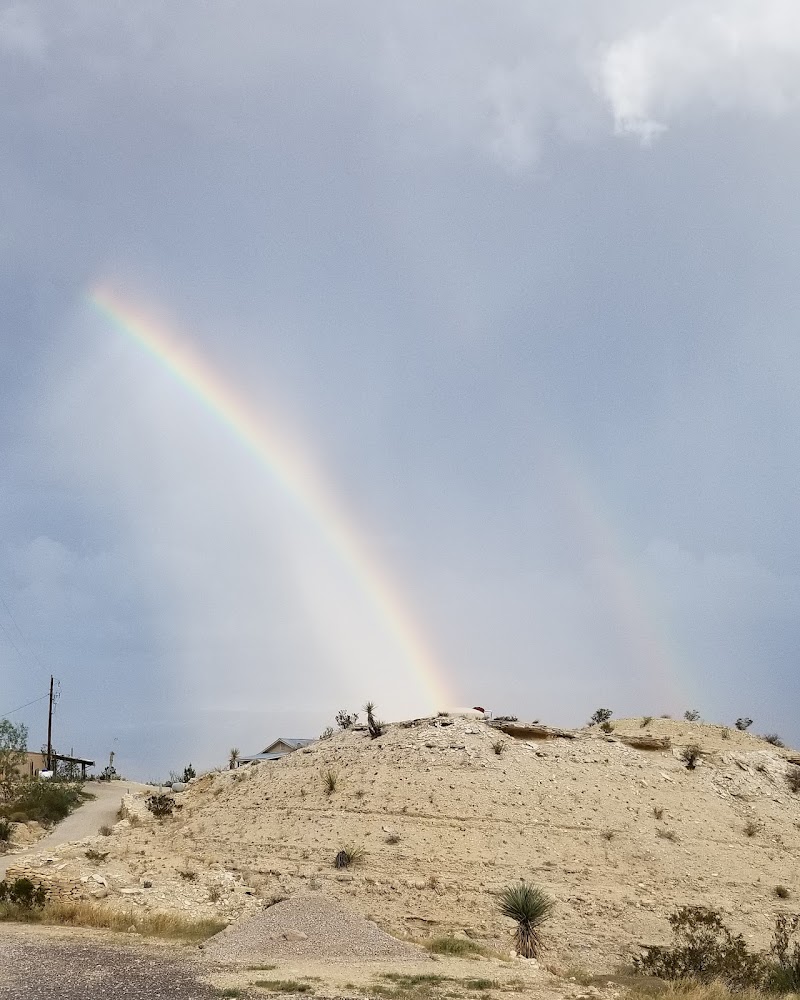 Rainbow arcs over Rancho Topanga Campground in Big Bend National Park, with a desert hill and scattered clouds in the background.