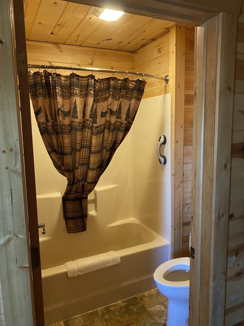 Rustic cabin bathroom at Badlands National Park with beige tub, plaid shower curtain, and a toilet beside wood walls.