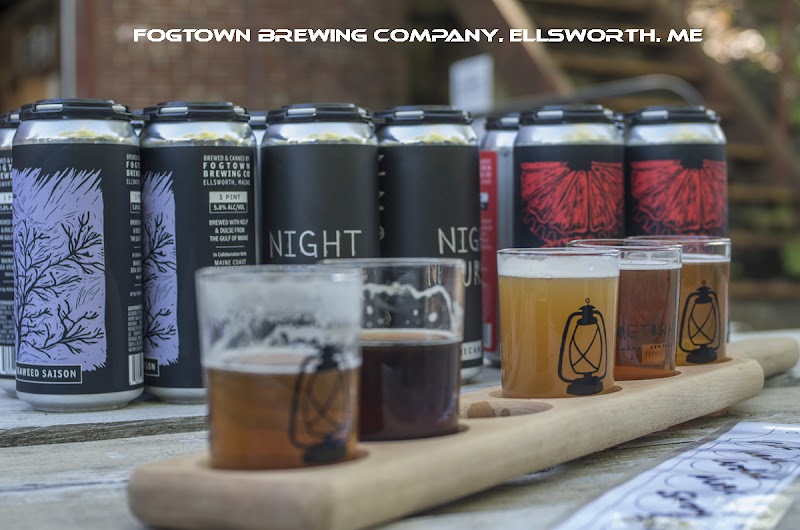 Tasting flight of beers displayed on a wooden board near Acadia National Park in Ellsworth, with colorful cans in the background.