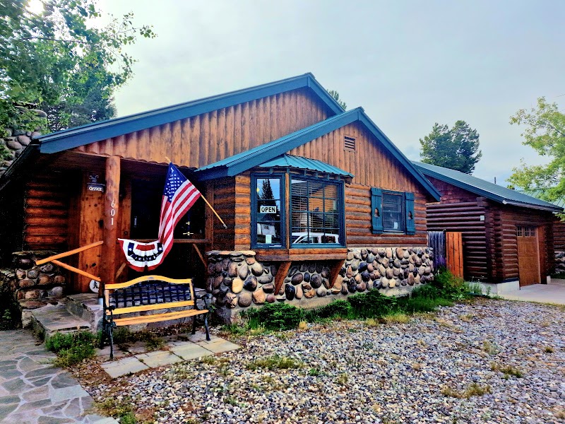 Cozy log inn with stone base, American flag, bench, open sign, and gravel lot in Yellowstone National Park.