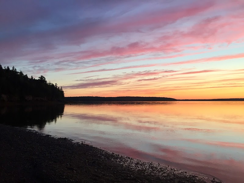 Sunset over a calm inlet with pink-orange sky, forested shoreline left, and mirror-like water at Acadia National Park.