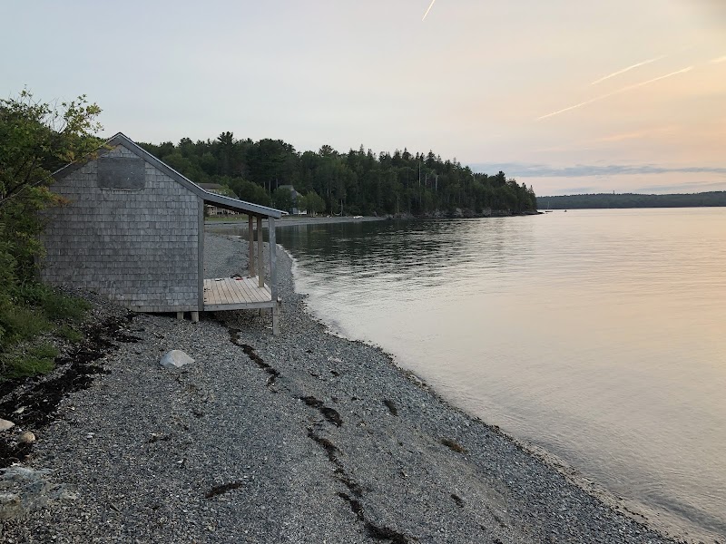 Shingled lakeside cottage on a pebbled shore with calm waters, forested shoreline, and pink sunset in Acadia National Park.