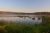 Mist over a calm lake with grassy islands, foreground reeds, and a pine shoreline at sunrise in Acadia National Park.