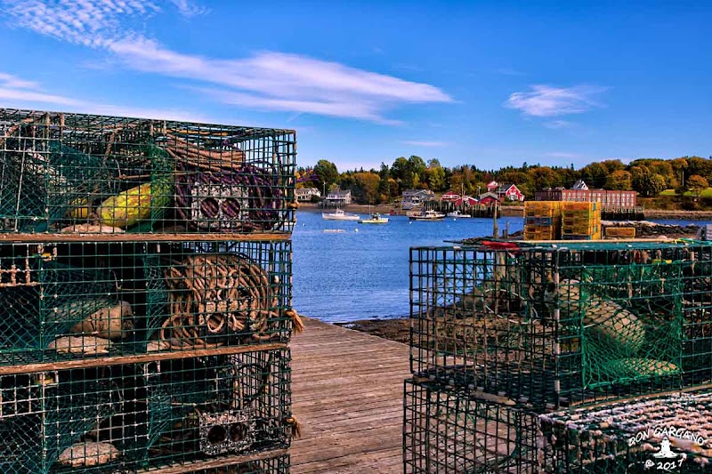 Lobster traps in wire cages stacked on a wooden dock overlook Bass Harbor in Acadia National Park, with blue harbor water and distant boats.