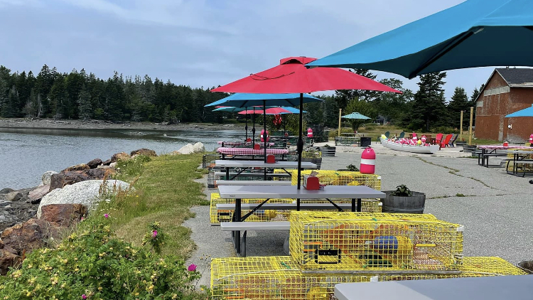 Outdoor dining area along a rocky shoreline with colorful umbrellas, tables, and yellow lobster traps at Acadia National Park.