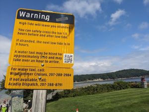 Bright yellow warning sign about tides near the Bar Island Land Bridge trail in Acadia National Park, with water, trees, and a grassy overlook.