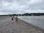 Hikers walk along a pebbled beach on the Bar Island Land Bridge in Acadia National Park, with calm water to the right and distant hills under cloudy skies.