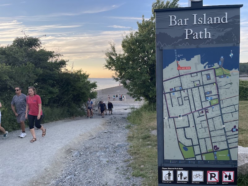 Gravel path toward a rocky shore at sunset in Acadia National Park, with a Bar Island Path map sign on the right and hikers.