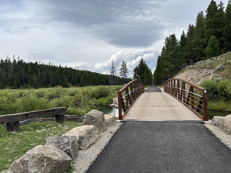 Wooden arch bridge spans a paved trail through a grassy meadow and pine forest in Yellowstone National Park.