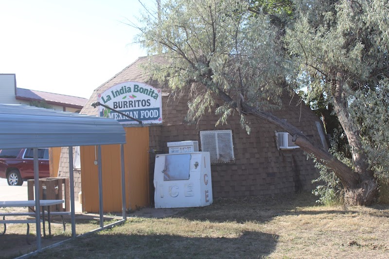 A small brown shingle building with a weathered sign, leaning tree, ice chest, and metal canopy in Badlands National Park.