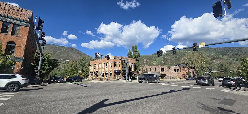 Busy street intersection with red brick buildings, cars waiting at a crosswalk, traffic signals overhead, and green hills under a blue sky in Badlands National Park.