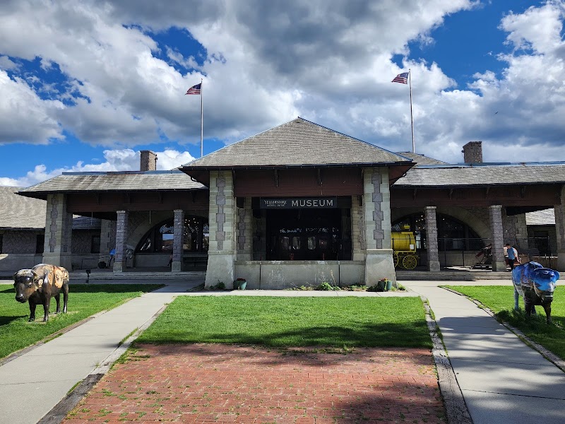 Stone-pillared museum in West Yellowstone with green lawn, brick path, bison statues, and flags, Yellowstone National Park.