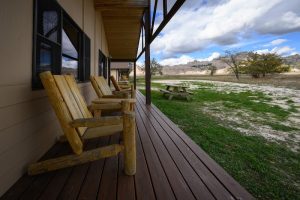 Wooden porch with rustic log chairs along a beige building, a picnic table on grass, with Badlands National Park in the distance.
