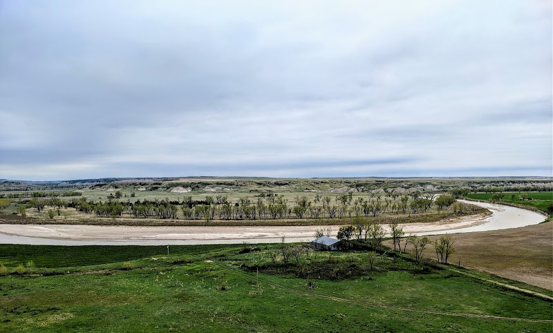 Winding gravel road snakes through grassy Badlands National Park, with a small white shelter and sparse trees under a cloudy sky.
