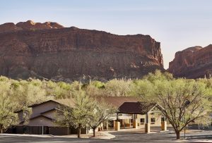 Lodging complex with tan buildings and green trees in front of red rock cliffs at Arches National Park.