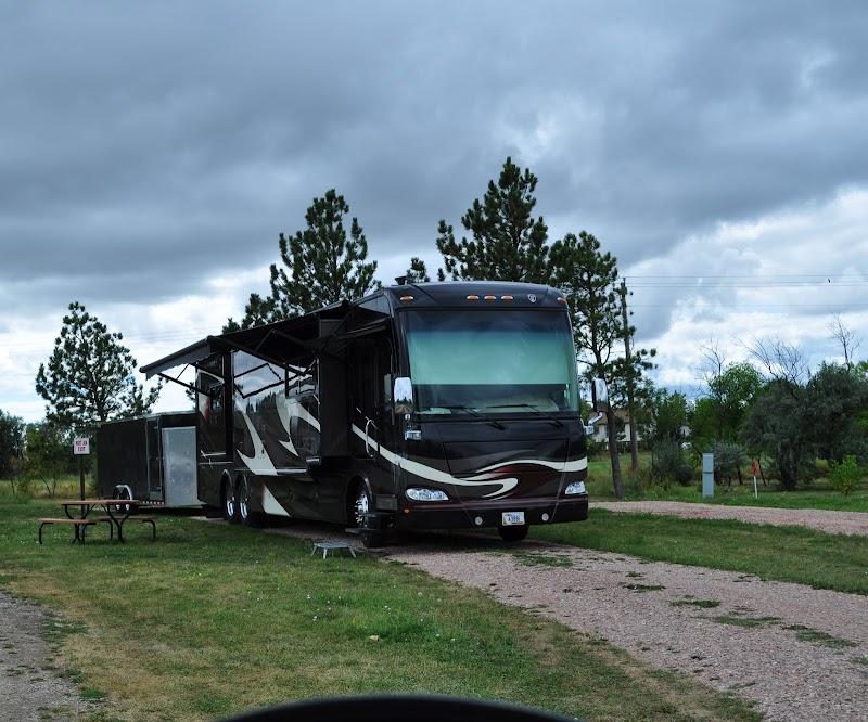Large dark RV with extended slides parked on a gravel loop at Badlands National Park campground, under a cloudy sky with a nearby picnic table.