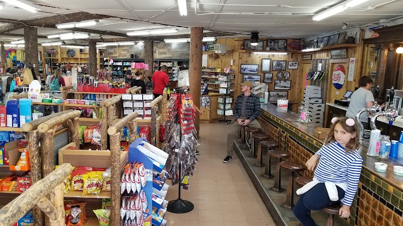 Inside a rustic Yellowstone National Park gift shop, shelves brim with snacks and souvenirs, while a girl in a striped shirt sits near a long counter.