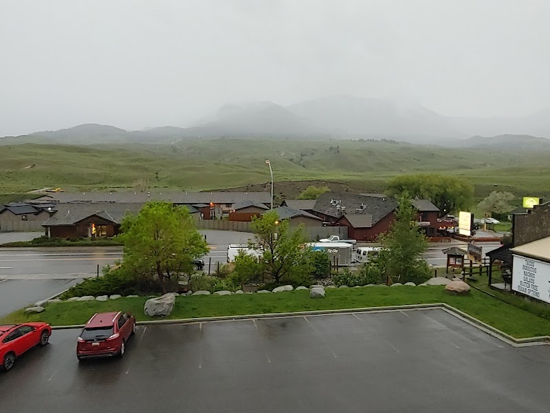 Low wooden lodge cabins line a wet road with parked cars, green shrubs, and misty hills in Yellowstone National Park.