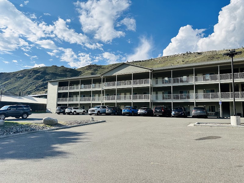 Three-story motel with balconies and a line of parked cars, set against green hills and a blue sky in Yellowstone National Park.