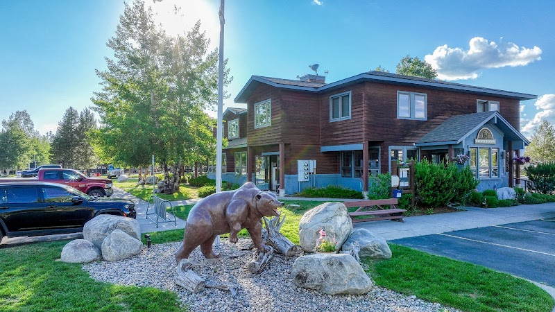Wooden visitor building beside a bear statue on rocks, with cars, trees, and gravel in Yellowstone National Park.