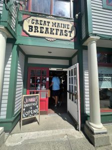 Entrance of a classic storefront with green trim and red doors, chalkboard sign outside, open doors, in Acadia National Park.