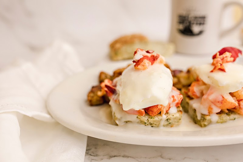 Plate of toast bites topped with poached eggs, creamy hollandaise, and pink lobster chunks, with a mug in the background at Acadia National Park.