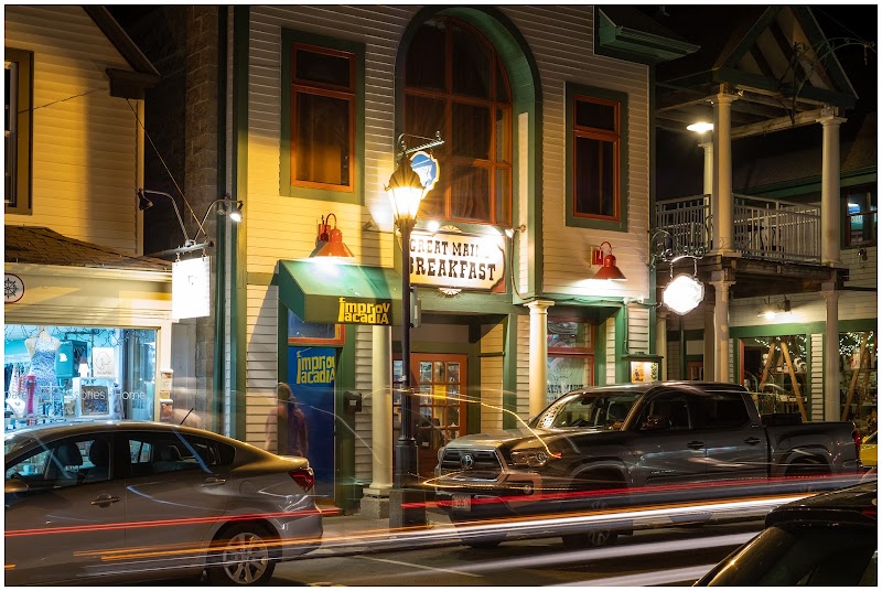 Nighttime street scene in Acadia National Park with a two-story diner-style storefront, green trim, arched window, glowing sign, and parked cars.