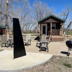 Patio chairs encircle a small wooden cabin with a green roof at Badlands National Park, beside a tall black obelisk sculpture.