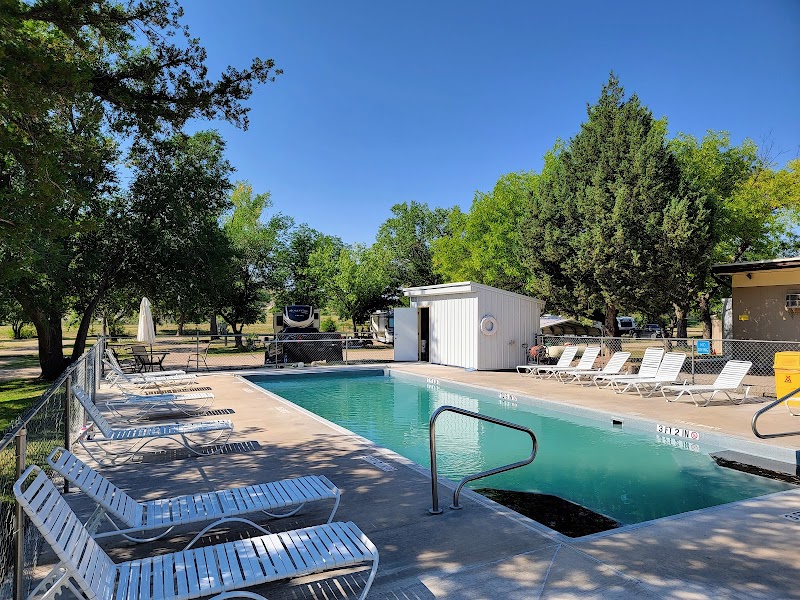Sunlit pool area at Badlands National Park campground with white lounge chairs, a small white pool building, and green trees under a clear blue sky.