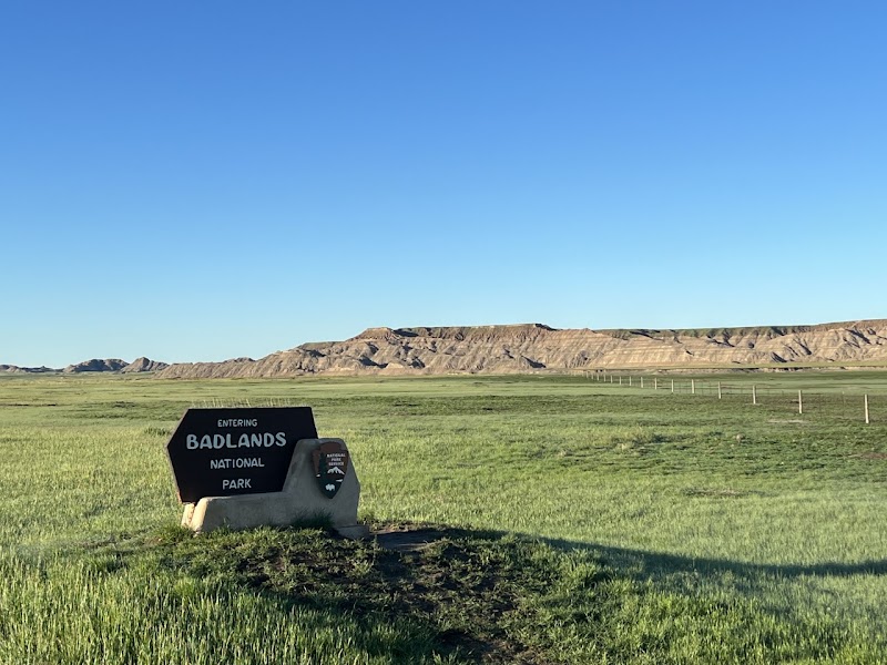 Open grassy plain under a clear blue sky with distant layered badlands and an 'Entering Badlands National Park' sign.