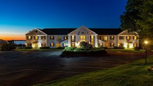 Lodge-style white buildings with glowing windows surround a parking lot, shrubs, and a sunset sky in Acadia National Park.
