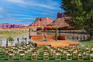 Outdoor wedding setup with wooden chairs facing a rustic ceremony platform by a river, framed by red rock mesas in Arches National Park.
