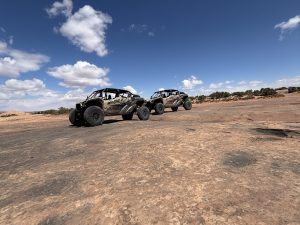 Two off-road rock crawlers parked on a wide reddish desert slab under a bright blue sky in Arches National Park.
