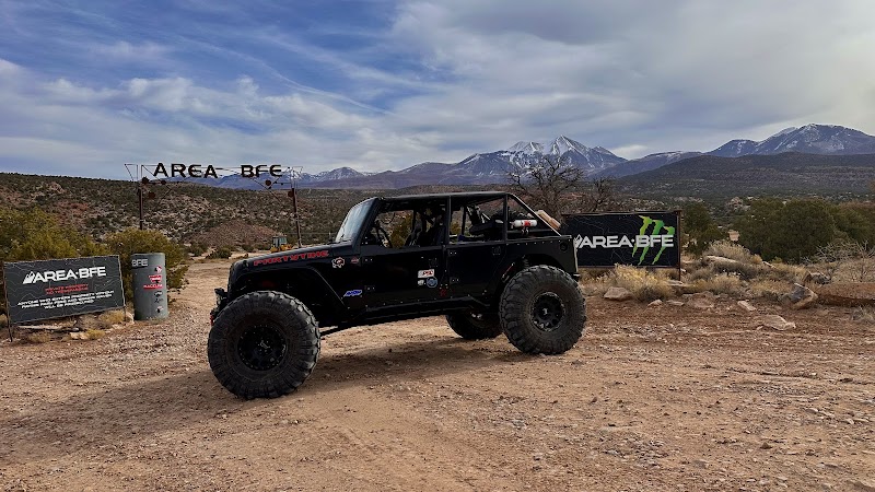 Black off-road Jeep with oversized tires on a dirt lot near a sign in Arches National Park, with desert shrubs and snow-capped mountains in the background.