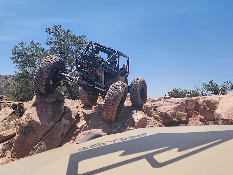 Off-road rock crawler tilts over large boulders at AreaBFE campground in Arches National Park under a clear blue sky.