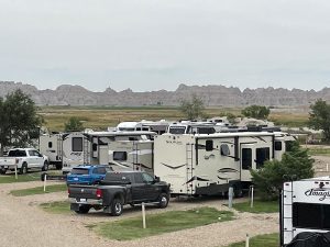 RVs, trucks, and campers parked at a Badlands National Park campground with distant rugged badlands.