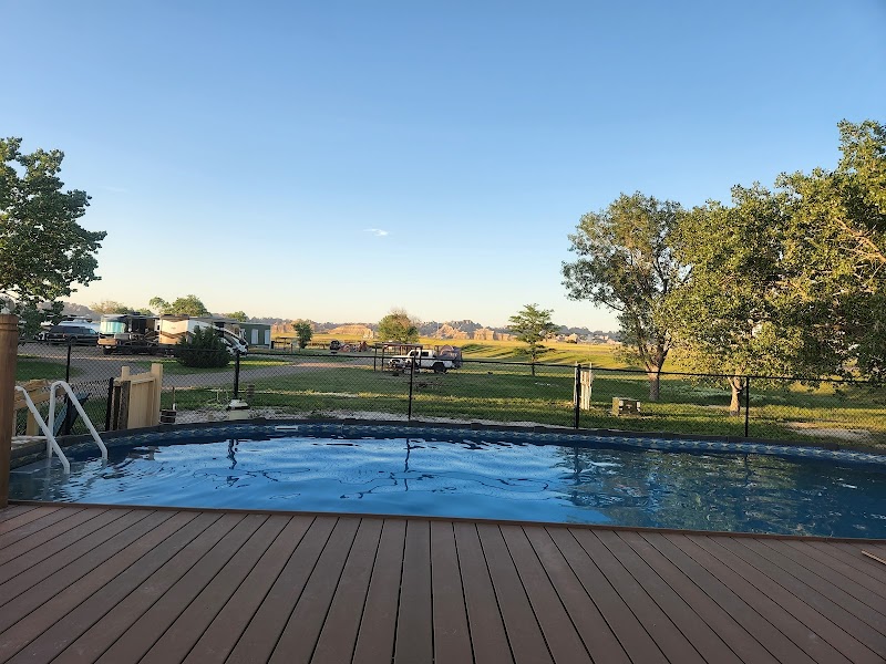 Wooden deck pool beside a grassy campground with RVs, trees, and a distant fence in Badlands National Park.