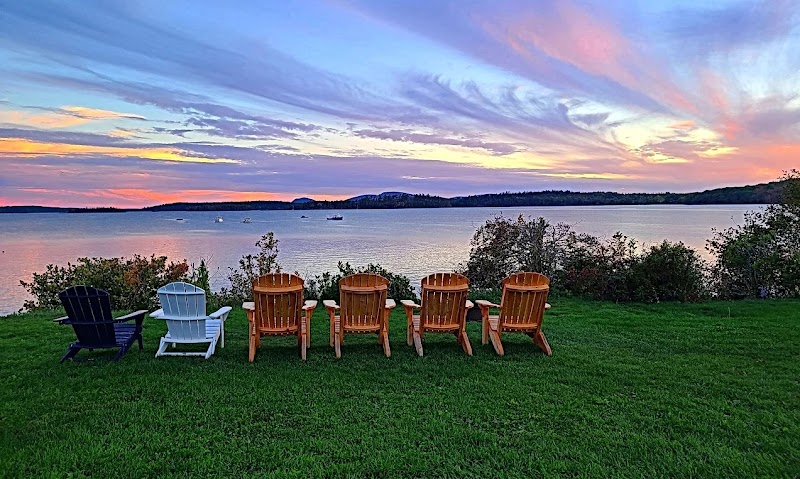 Sunset over a calm bay in Acadia National Park with seven Adirondack chairs lined up on green grass facing the water.