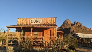Rustic wooden storefront with a 'GUNS' sign sits in a sunlit desert at Big Bend National Park.